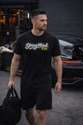 Man wearing a black 'StrongMind Co.' t-shirt walking outdoors with a car in the background.