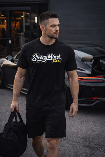 Man wearing a black 'StrongMind Co.' t-shirt walking outdoors with a car in the background.